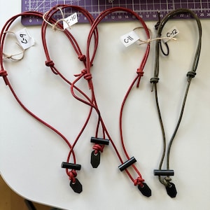 May include: Four leather cord necklaces in red and olive green. Each necklace has a black rectangular bead and a black tag. The necklaces are labeled with tags that read "6-7", "C-10", "C-11", and "C-12".
