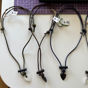 May include: A collection of handmade necklaces with arrowhead pendants. The necklaces feature dark cords and black cylindrical beads. The pendants are in various colors, including white and black. Each necklace has a tag with a handwritten number.