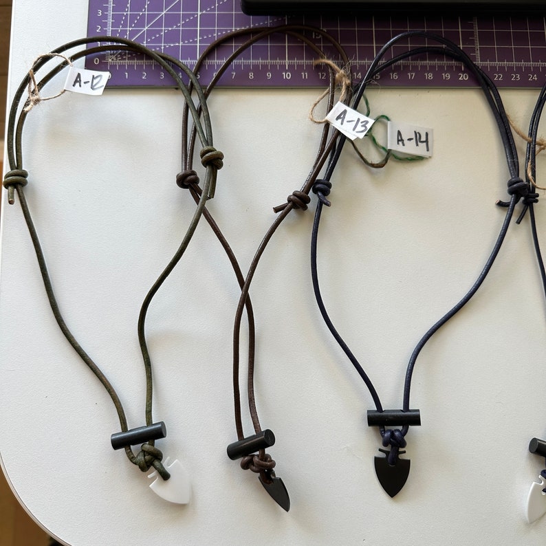 May include: A collection of handmade necklaces with arrowhead pendants. The necklaces feature dark cords and black cylindrical beads. The pendants are in various colors, including white and black. Each necklace has a tag with a handwritten number.