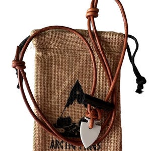 May include: A brown leather necklace with a white arrowhead pendant and a black cylindrical accent. The necklace is displayed in front of a burlap drawstring bag with a logo and text. The bag is tan in color.