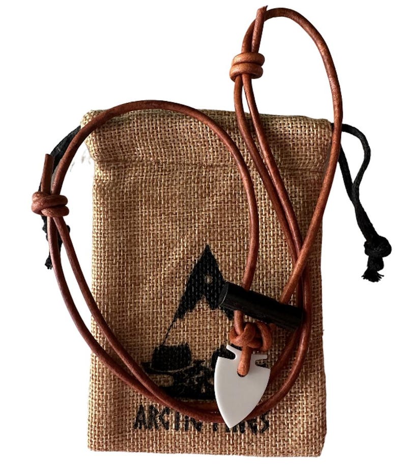 May include: A brown leather necklace with a white arrowhead pendant and a black cylindrical accent. The necklace is displayed in front of a burlap drawstring bag with a logo and text. The bag is tan in color.