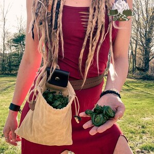 Könnte beinhalten: Eine Frau trägt ein rotes Kleid mit einem braunen Ledergürtel und eine beige Canvas-Tasche mit einer Klappe und einer Schnalle. Die Tasche ist mit grünen Geißblatt gefüllt. Die Frau hält eine Handvoll Geißblatt in ihrer rechten Hand. Der Text "ARCTIC PINES" ist in der oberen rechten Ecke des Bildes sichtbar.