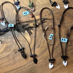 May include: A collection of handmade necklaces with arrowhead pendants. The necklaces feature leather cords in various colors, including black, brown, and purple. The pendants are made of different materials and colors, such as black, white, and silver. The necklaces are displayed on a wooden surface.