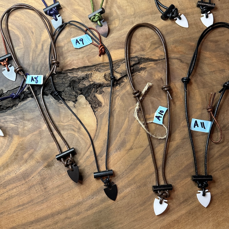 May include: A collection of handmade necklaces with arrowhead pendants. The necklaces feature leather cords in various colors, including black, brown, and purple. The pendants are made of different materials and colors, such as black, white, and silver. The necklaces are displayed on a wooden surface.