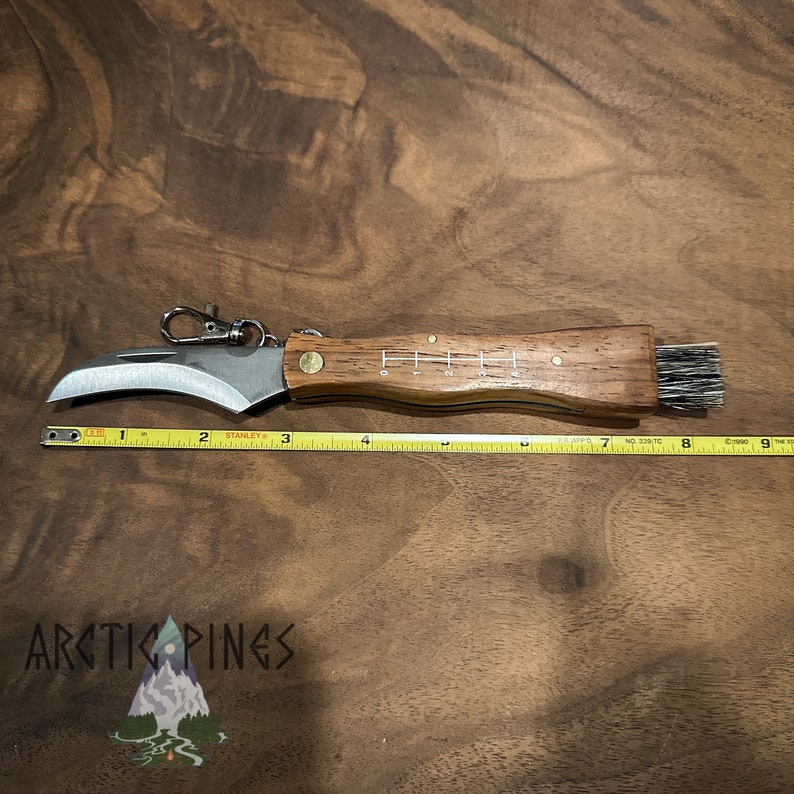 May include: A small, brown handled knife with a curved blade and a brush on the end of the handle. The knife is 7 inches long and has a key ring attached to the handle. The knife is sitting on a wooden surface.
