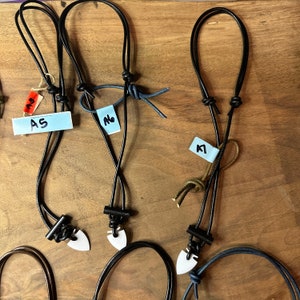 May include: Several leather necklaces with white arrowhead pendants are displayed on a wooden surface. The necklaces have black leather cords and are knotted at the top. Each necklace has a tag with a letter and number.