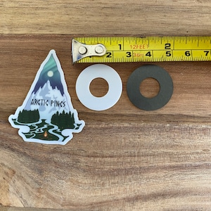 Könnte beinhalten: Ein Aufkleber mit einer Bergszene mit einem Lagerfeuer und dem Text "Arctic Pines". Der Aufkleber befindet sich auf einer Holzoberfläche mit einem weißen und schwarzen Kreis daneben.