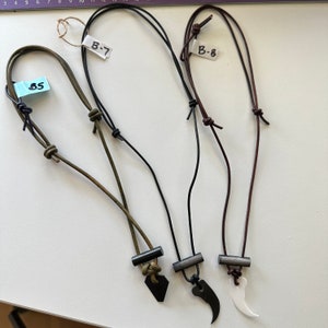 May include: Three leather cord necklaces with different pendant shapes. One has a black, pointed pendant, another a claw-shaped pendant, and the last a white, curved pendant. Each necklace has a metal tube accent and a tag with a number.