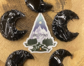 May include: Five crescent-shaped, polished black obsidian stones arranged on a wooden surface. A sticker with the text "ARCTIC PINES" and a mountain scene is in the center. The obsidian pieces have a glossy, reflective surface.