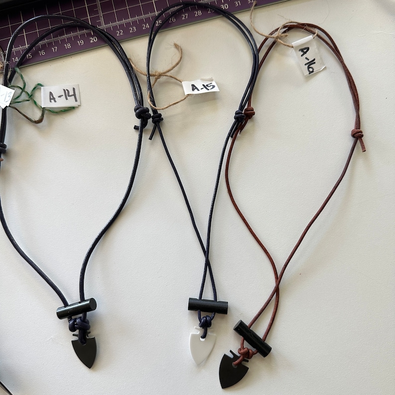 May include: Three handmade necklaces with arrowhead pendants. One necklace has a black pendant, one has a white pendant, and one has a black pendant. The cords are black, navy blue, and brown. Each necklace has a tag with a handwritten number.