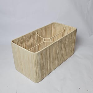 Peut inclure: Abat-jour rectangulaire en tissu beige naturel et texturé. Il possède une armature métallique et une forme rectangulaire arrondie. Le tissu présente des lignes verticales, donnant un aspect tissé. Conçu pour diffuser la lumière, il est un élément de décoration.