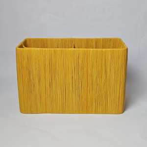 May include: A rectangular, yellow woven basket with a textured surface. The basket has a square opening and is divided into two compartments. The basket is made of a natural fiber material.