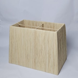 May include: Rectangular lampshade in a natural, textured finish. The light beige shade has a trapezoidal shape and is made from woven strands, giving it a rustic look. The lampshade has a metal frame. The dimensions are approximately 30 cm in height.