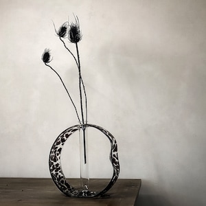 以下が含まれることがあります： 黒と白のパターンが施されたガラスの花瓶。丸い開口部と透明なガラスのチューブが内側にあります。乾燥した黒いアザミの花が花瓶に飾られています。