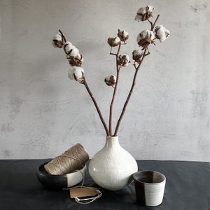 May include: A white ceramic vase with a textured surface holds a bouquet of dried cotton stems. The vase is sitting on a black surface with a spool of twine and a small ceramic cup.