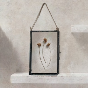 Op de afbeelding: Een zwart metalen frame met een touwbevestiging, met gedroogde bloemen erin. Het frame hangt aan een wit rek.