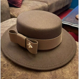 Könnte beinhalten: Ein brauner Filzhut im Fedora-Stil mit einem braunen Grosgrainband und einer goldenen Bienenbrosche. Der Hut hat eine breite Krempe und eine leicht abgerundete Krone.