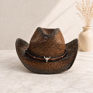 Può includere: Un cappello da cowboy marrone con un design intrecciato. Presenta una fascia in pelle con un emblema a teschio di toro argentato. La tesa è leggermente curva, su sfondo neutro.