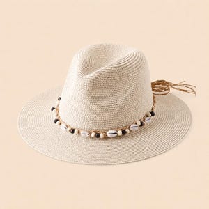Może przedstawiać: Kremowy kapelusz przeciwsłoneczny w stylu fedora z szerokim rondem. Kapelusz jest ozdobiony opaską z muszli i ciemnych drewnianych koralików. Z boku wisi ozdobny chwost. Kapelusz jest wykonany z plecionej słomy.