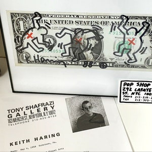 Puede incluir: Un billete de un d&oacute;lar enmarcado con un dibujo en blanco y negro de figuras de palo. El dibujo est&aacute; firmado "Keith Haring". El marco es negro y el paspart&uacute; es blanco. Hay una tarjeta de visita debajo del marco que dice "Pop Shop 292 Lafayette St. NYC 10012 Phone 212-219-2784 Fax 212-353-4843".