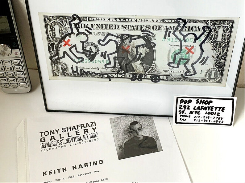 Puede incluir: Un billete de un d&oacute;lar enmarcado con un dibujo en blanco y negro de figuras de palo. El dibujo est&aacute; firmado "Keith Haring". El marco es negro y el paspart&uacute; es blanco. Hay una tarjeta de visita debajo del marco que dice "Pop Shop 292 Lafayette St. NYC 10012 Phone 212-219-2784 Fax 212-353-4843".