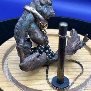 Puede incluir: Una escultura de tono bronce con un acabado texturizado y desgastado, que presenta una cola prominente y apéndices en forma de garra. La figura sostiene un grupo de pequeñas cuentas metálicas oscuras y lleva un anillo circular de tono dorado.