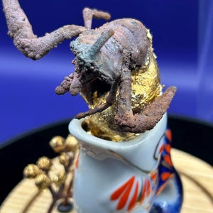 Puede incluir: Una escultura texturizada de color marrón que recuerda a un crustáceo emerge de un recipiente de cerámica blanca con detalles pintados en naranja y azul. La escultura está parcialmente cubierta de pan de oro y una rama con bayas doradas.