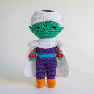 Puede incluir: Una figura amigurumi de crochet de un personaje de piel verde con un turbante blanco, un atuendo morado y botas naranjas. El personaje tiene una expresión seria y está de pie sobre un fondo blanco.