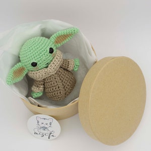 Peut inclure: Un jouet en peluche crochet&eacute; vert et marron en forme de personnage de la franchise Star Wars, assis dans une bo&icirc;te cadeau ronde et brune avec un rembourrage en papier de soie blanc. La bo&icirc;te a un autocollant avec une illustration de chat et le texte "mischief".