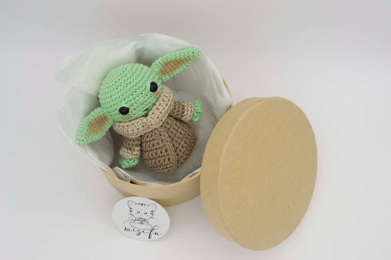 Peut inclure: Un jouet en peluche crochet&eacute; vert et marron en forme de personnage de la franchise Star Wars, assis dans une bo&icirc;te cadeau ronde et brune avec un rembourrage en papier de soie blanc. La bo&icirc;te a un autocollant avec une illustration de chat et le texte "mischief".