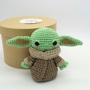 Peut inclure: Un jouet en peluche crochet&eacute; vert et marron ressemblant &agrave; un personnage de la franchise Star Wars. Le jouet a de grands yeux et une petite bouche. Il porte un manteau crochet&eacute; marron.