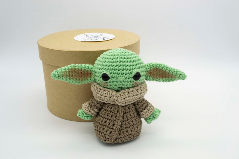 Peut inclure: Un jouet en peluche crochet&eacute; vert et marron ressemblant &agrave; un personnage de la franchise Star Wars. Le jouet a de grands yeux et une petite bouche. Il porte un manteau crochet&eacute; marron.