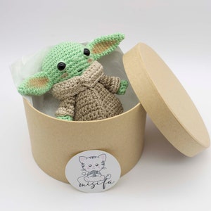 Peut inclure: Un jouet en peluche crochet&eacute; vert et marron en forme de personnage de la franchise Star Wars, assis dans une bo&icirc;te en carton marron avec un couvercle. La bo&icirc;te a un autocollant blanc avec un chat de dessin anim&eacute; et le texte "misifu".
