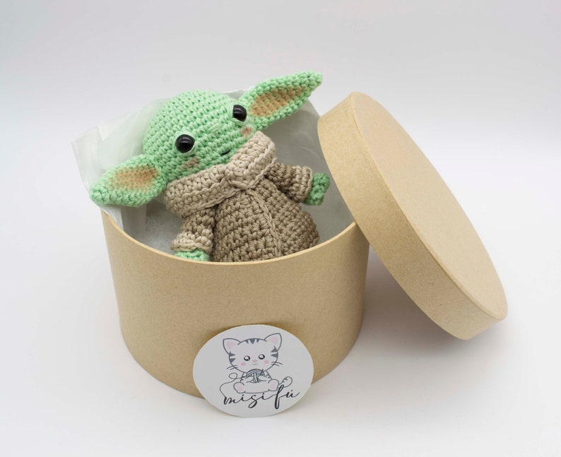 Peut inclure: Un jouet en peluche crochet&eacute; vert et marron en forme de personnage de la franchise Star Wars, assis dans une bo&icirc;te en carton marron avec un couvercle. La bo&icirc;te a un autocollant blanc avec un chat de dessin anim&eacute; et le texte "misifu".