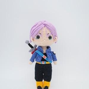Puede incluir: Una figura de crochet de un personaje con cabello morado, una chaqueta azul, pantalones negros y zapatos amarillos. La figura sostiene una espada.