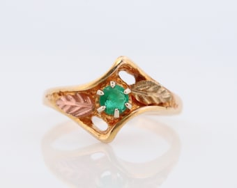 May include: A gold ring featuring a green emerald gemstone set within a leaf design. The ring has a delicate, feminine style.