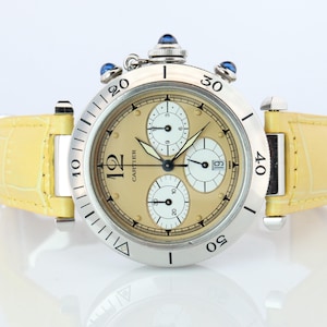 May include: A Cartier wristwatch featuring a round silver-tone stainless steel case with a rotating bezel, a beige dial with three white sub-dials, a date window, and a yellow textured leather strap with a prominent crown topped with a blue cabochon.