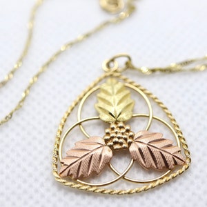 May include: A gold-toned necklace with a pendant featuring a leaf design. The pendant has three leaves in gold and rose gold, with a cluster of small gold beads in the center. The chain is a delicate, twisted design.