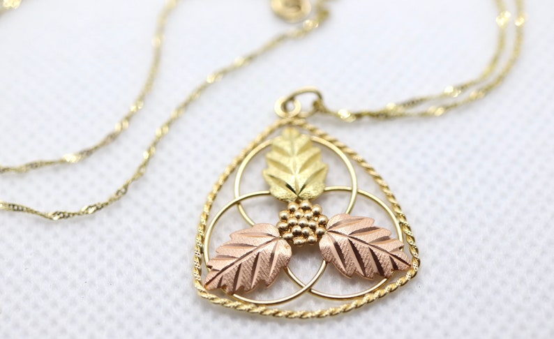 May include: A gold-toned necklace with a pendant featuring a leaf design. The pendant has three leaves in gold and rose gold, with a cluster of small gold beads in the center. The chain is a delicate, twisted design.