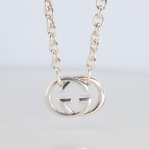 Puede incluir: Collar plateado con un colgante doble G. El colgante presenta el logotipo GG entrelazado en un diseño circular. La cadena es de estilo eslabón delicado. El collar se presenta sobre un fondo blanco.