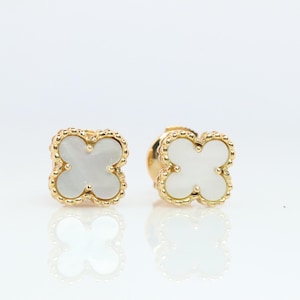 May include: A pair of gold-colored stud earrings, each featuring a four-leaf clover design with a white, pearlescent center. The earrings are set against a white background, with their reflections visible below.