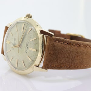 Pu&ograve; includere: Un orologio da polso dorato con un cinturino in pelle marrone. Il quadrante dell'orologio &egrave; bianco con segni neri e la scritta "21 Jewels Automatic" stampata su di esso.