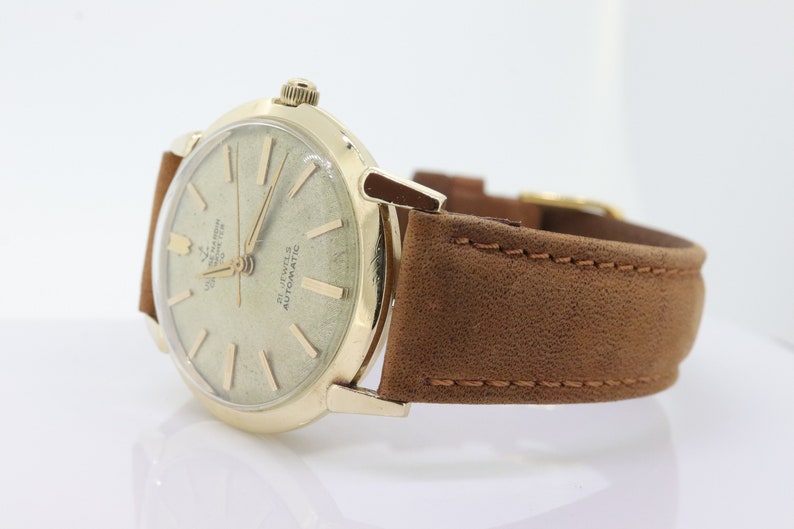 Pu&ograve; includere: Un orologio da polso dorato con un cinturino in pelle marrone. Il quadrante dell'orologio &egrave; bianco con segni neri e la scritta "21 Jewels Automatic" stampata su di esso.