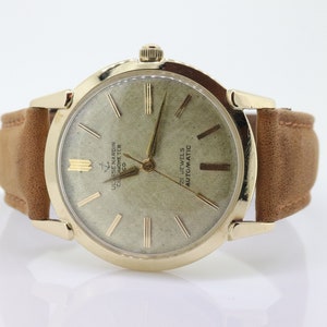 Pu&ograve; includere: Un orologio da polso dorato con quadrante bianco e lancette nere. L'orologio ha un cinturino in pelle marrone e il testo "ULYSSE NARDIN CHRONOMETER  CHICAGO  CO" e "21 JEWELS AUTOMATIC" stampato sul quadrante.