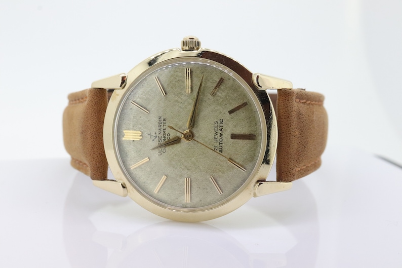 Pu&ograve; includere: Un orologio da polso dorato con quadrante bianco e lancette nere. L'orologio ha un cinturino in pelle marrone e il testo "ULYSSE NARDIN CHRONOMETER  CHICAGO  CO" e "21 JEWELS AUTOMATIC" stampato sul quadrante.