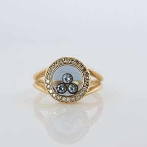 May include: A gold ring featuring a circular centrepiece with a clear glass window. Inside the window are three small, loose, round, light blue stones. The outer edge of the circular frame is set with a border of tiny, sparkling white diamonds.