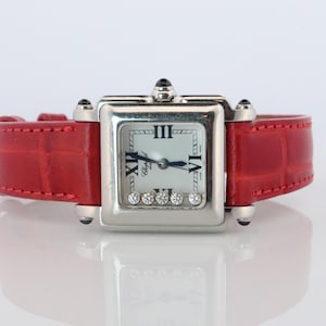 May include: A square-faced wristwatch with a silver-tone metal case and a bright red leather strap featuring a textured finish. The white dial displays black Roman numerals and five small, loose round diamonds positioned at the bottom.