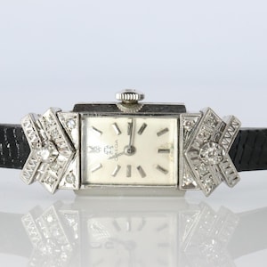 May include: A silver-toned Omega wristwatch featuring a rectangular white dial with silver hour markers and hands. The watch case is flanked by geometric silver attachments set with small clear stones, connected to a black textured leather strap.