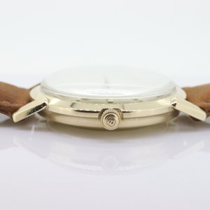 Pu&ograve; includere: Un orologio da polso dorato con quadrante bianco e cinturino in pelle marrone. L'orologio ha un design semplice con una sola lancetta e una piccola corona sul lato.