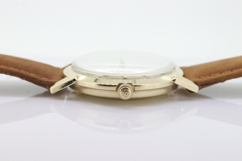 Pu&ograve; includere: Un orologio da polso dorato con quadrante bianco e cinturino in pelle marrone. L'orologio ha un design semplice con una sola lancetta e una piccola corona sul lato.